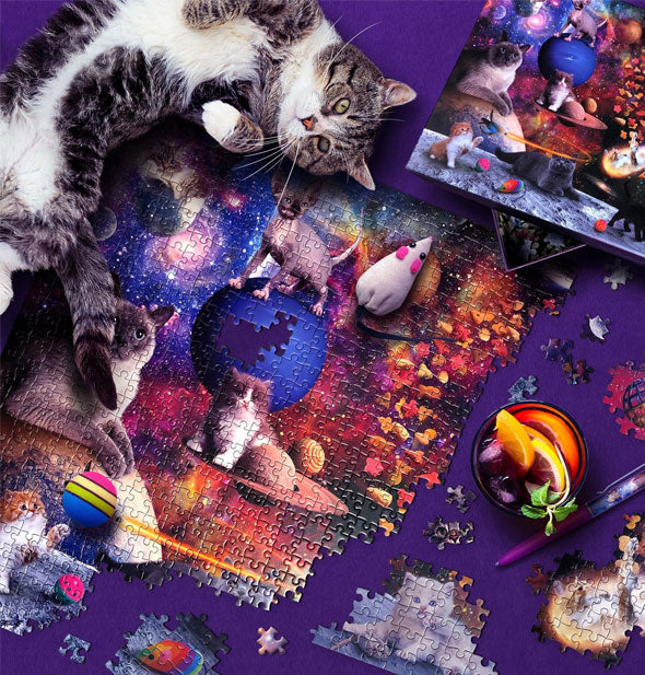 A grey and white tabby sprawls across a mostly assembled Galaxy Cats jigsaw puzzle with cocktail, pen, and loose pieces nearby