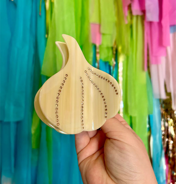 Model's hand holds a garlic bulb hair clip with rhinestone details in front of a colorful streamer backdrop