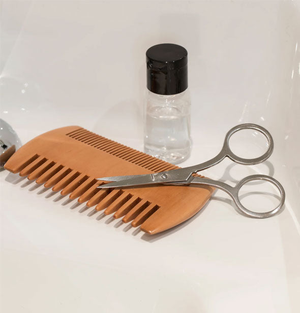 Double-sided wooden beard comb, stainless steel scissors, and oil bottle rest on a sink basin's edge