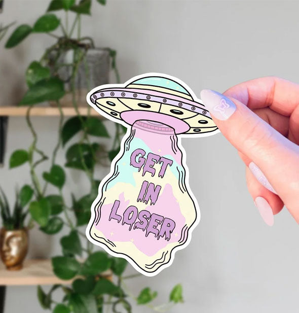 Model's hand holds a pastel blue, yellow, and purple spaceship sticker with the words, "Get in loser" printed inside its beam