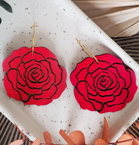 Red rose earrings with engraved black petal details hang from gold hoops and rest on a white speckled dish