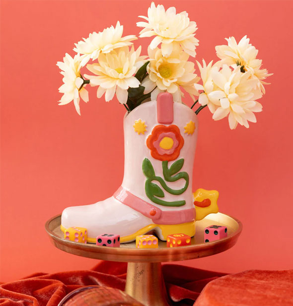 Cowgirl boot flower vase holds a small bouquet and rests on a gold pedestal surrounded by colorful dice against a coral backdrop