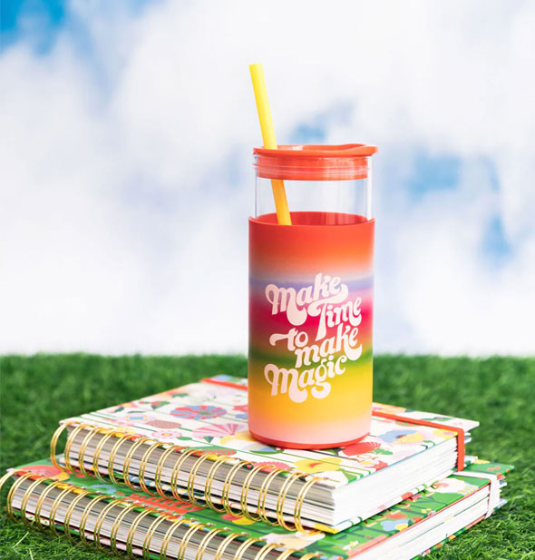 Make Time to Make Magic drink tumbler on top of two stacked spiral-bound notebooks on green astroturf against a blue sky with white clouds backdrop