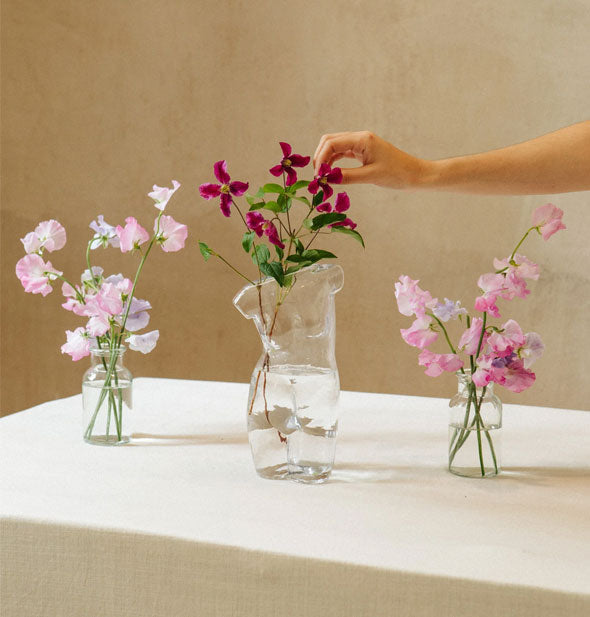 Clear glass nude form vase holds dark purple flowers being adjusted by a model's hand on a tabletop with two other flower-bearing vases