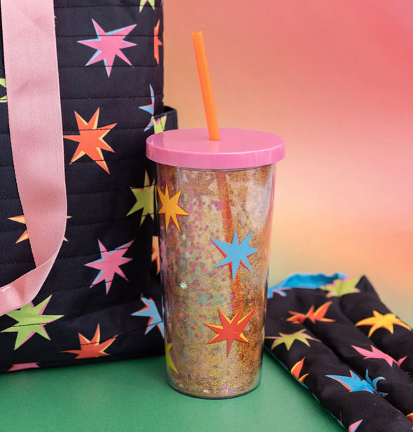 Glitter Starburst tumbler staged on a green surface with starburst-patterned cloth bags against a pink-to-orange ombre backdrop