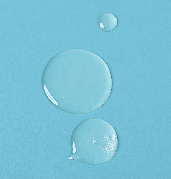 Closeup of clear, slightly bubbly droplets of Verb Glossy Shampoo on a blue surface