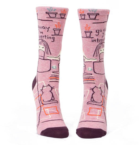 Pair of pink crew socks with dark toes features embroidered design of girl sipping from a mug with cat, books, and plants nearby