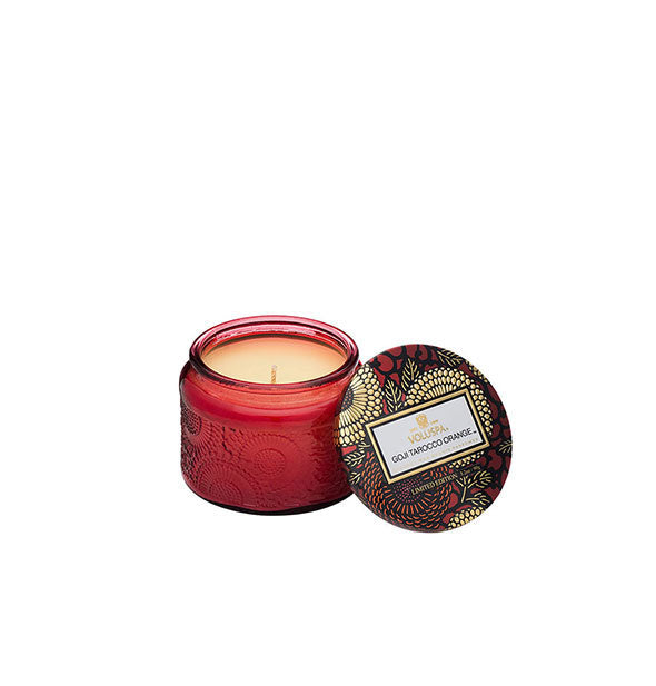 Red embossed glass Goji Tarocco Orange Voluspa candle har with red and gold metallic floral lid set to the side