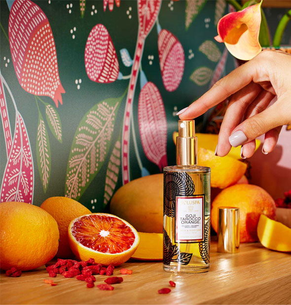 A model's finger presses down on the gold nozzle of a bottle of Voluspa Goji Tarocco Orange Room & Body Spray staged on a wooden surface with tropical fruits and citrus against a stylized pomegranate patterned backdrop