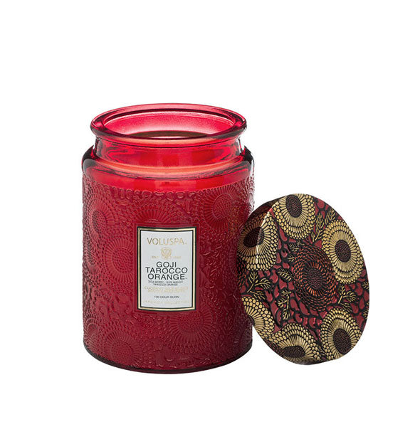 Red embossed glass Voluspa Goji Tarocco Orange candle jar with gold, red, and black floral patterned tin lid removed and propped up alongside