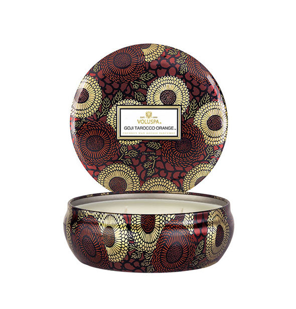 Round red, gold, and black floral patterned Voluspa Goji Tarocco Orange candle tin with lid removed and set upright behind the tin