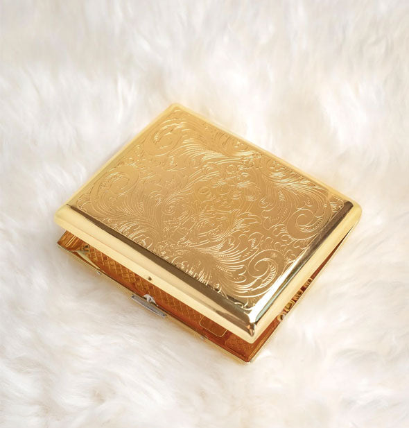 Rectangular gold cigarette case with ornate floral etching rests on white fur