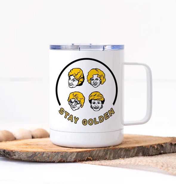 White mug with squared handle resting on a wooden slab features small portraits of the Golden Girls in a semicircle says, "Stay Golden"
