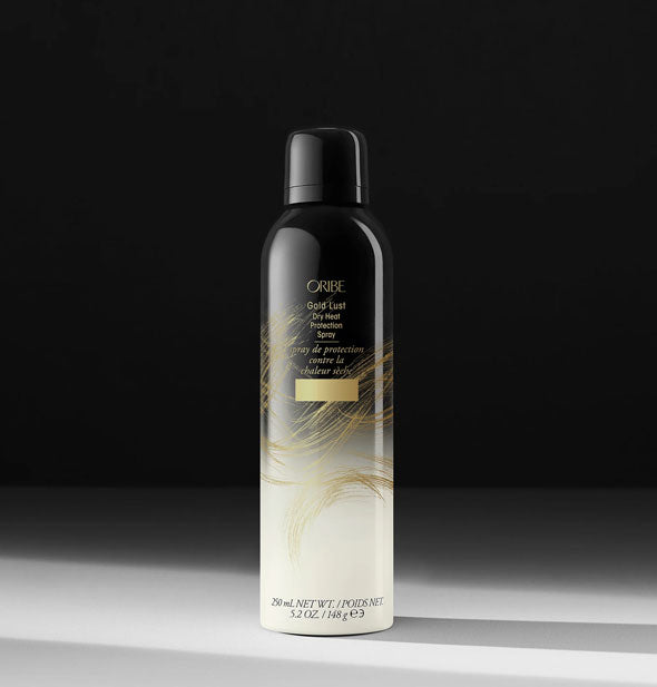 Black-to-white ombre 5.2 ounce can of Oribe Gold Lust Dry Heat Protection Spray with gold lettering and design accents on a heavily shadowed backdrop