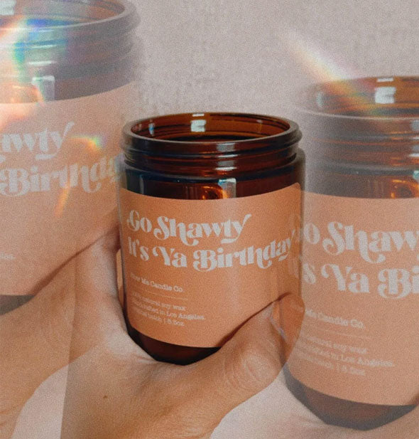 Repeated image of a model's hand holding am amber glass jar candle with peach-colored label that says, "Go Shawty It's Ya Birthday" in white lettering