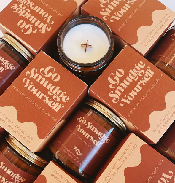 Assortment of Go Smudge Yourself candle jars and boxes; one candle's lid has been removed to reveal its wooden X-shaped wick