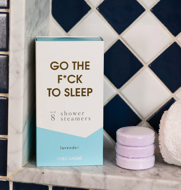 Go the F*ck to Sleep shower steamers box and three light purple steamer discs rest on a marble bath ledge next to a rolled-up white washcloth in front of black and white tile