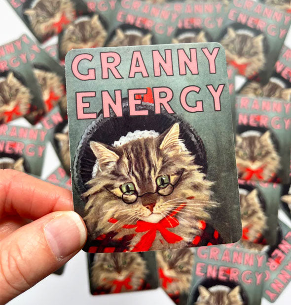 Model's hand holds a Granny Energy cat sticker in front of a pile of others like it in the background
