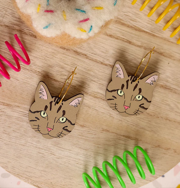 Pair of grayish-brown tabby cat faces on gold earring hoops rest on a wooden surface with brightly colored coils and a plush toy