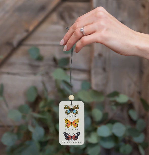 Model's hand holds a Grow Evolve Transform butterflies air freshener in front of a vine-covered wood backdrop