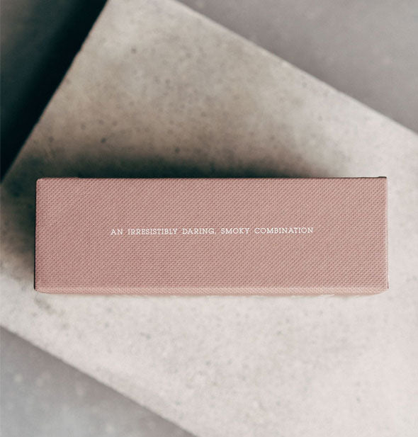 Textured blush rose rectangular box resting on a granite block is printed with small white lettering: "An irresistibly daring, smoky combination"