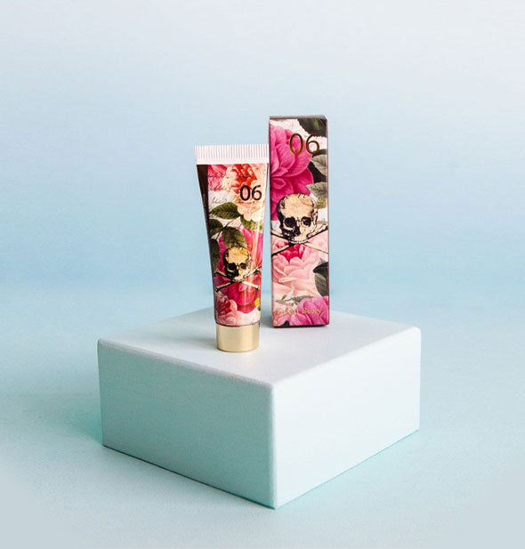 Tube and box of pink floral print handcreme with a skull and crossbones graphic at center of each rest on a small blue block