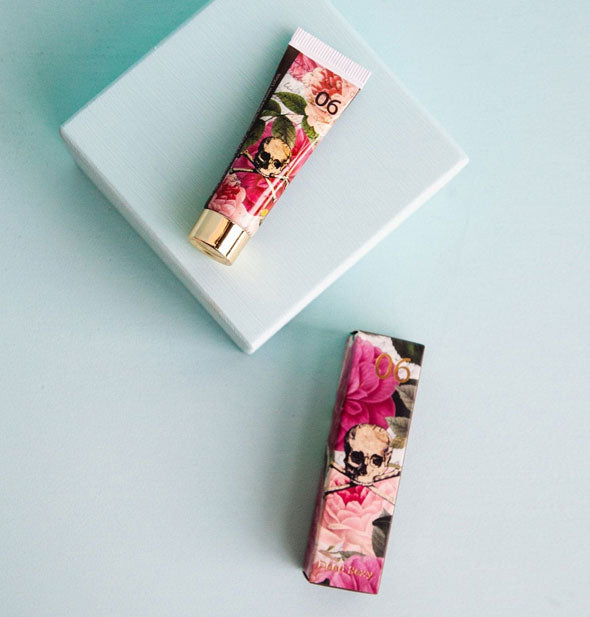Tube and box of pink floral print handcreme with a skull and crossbones graphic at center of each rest on and beside a small blue block