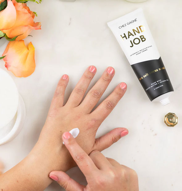 Model applies a small dollop of Hand Job hand cream to the back of left hand with right index finger; to the left are peach-colored roses