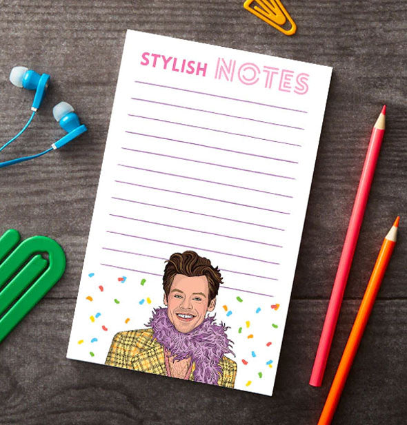 Stylish Notes Harry Styles notepad rests on a wooden surface with green and yellow paper clips, blue earbuds, and red and orange colored pencils