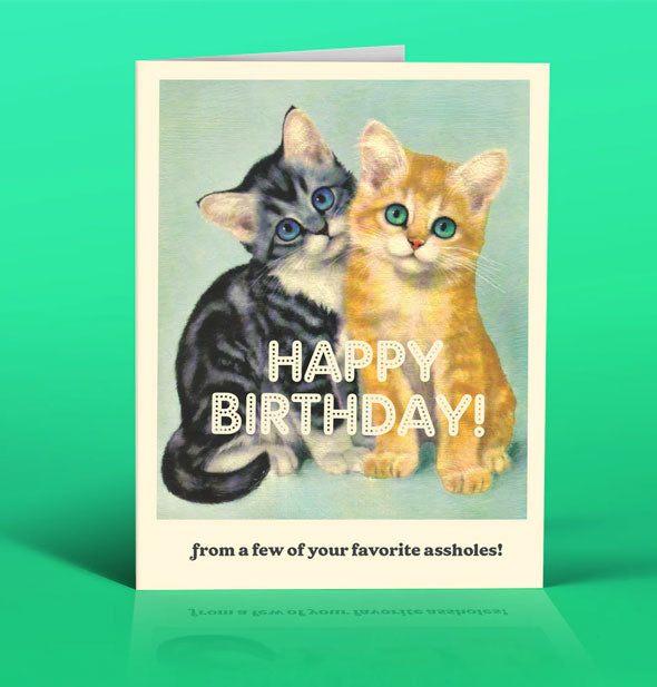 Greeting card on green backdrop features illustration of one black and one yellow kitten with the message, "Happy birthday! From a few of your favorite assholes!"