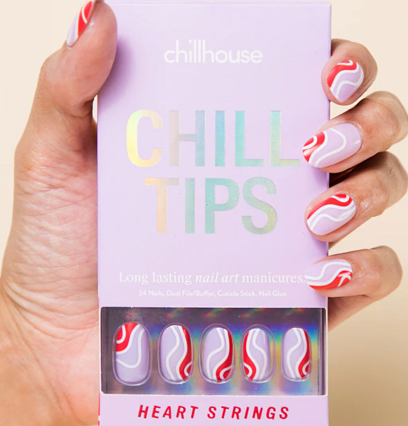 Model's hand wearing press-on nails in a purple and red pattern with wavy white line accents holds a box of Chillhoues Chill Tips Heart Strings nails of the same style