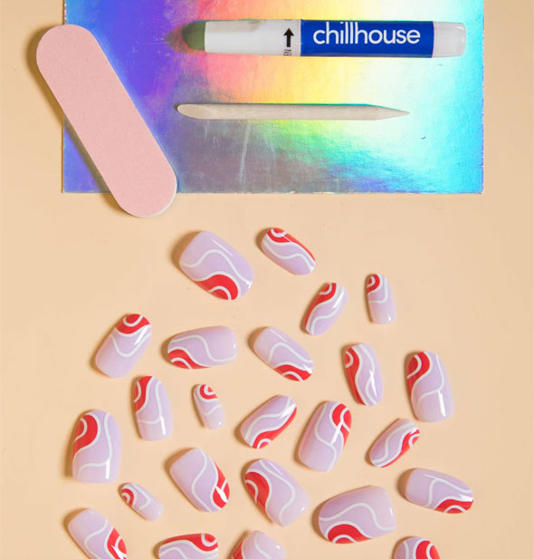 A smattering of red, purple, and white wavy line design press-on nails next to an iridescent tray holding Chillhouse glue tube, pink nail file, and wooden cuticle pusher
