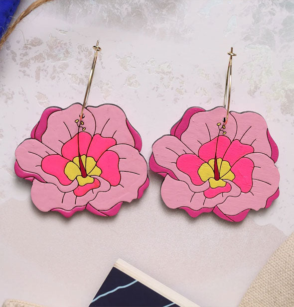 Pink and yellow hibiscus flowers on gold earring hoops rest on a concrete surface