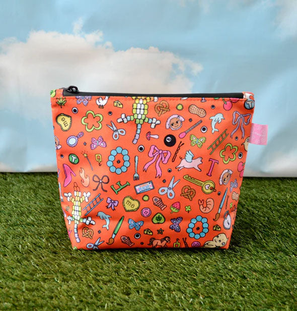 Orange pouch with black zipper and small illustrations of random objects all over it rests on green astroturf in front of a blue sky and clouds backdrop