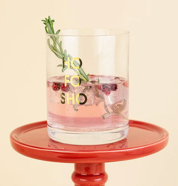 Ho Fo Sho rocks glass on red pedestal is filled with a bubbly pink beverage and garnished with pomegranate seeds and fresh herb sprig