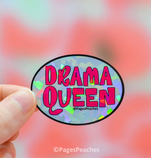 Model's hand holds an oval-shaped holographic sticker that says, "Drama queen" in large pink lettering inside a thin black border