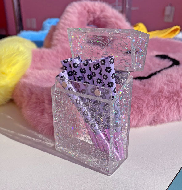 Clear cigarette box with holographic glitter flecks throughout holds several rolled cigarettes with purple daisy print paper