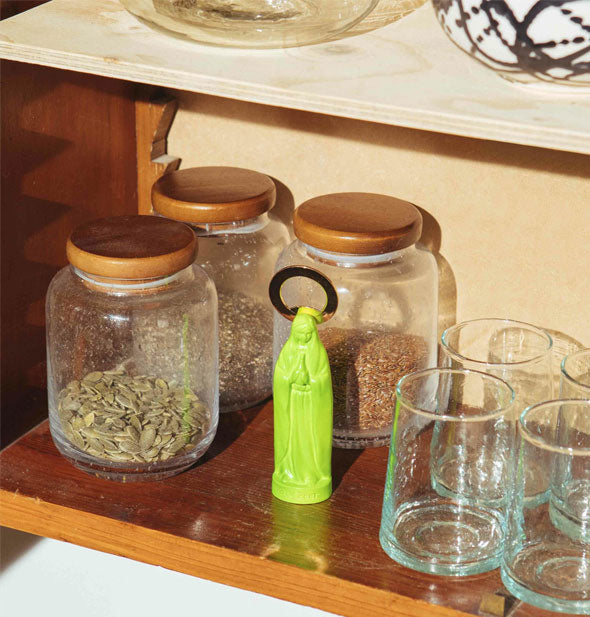 A green Holy Beer Bottle Opener sits on a wooden shelf with barware and jars of seeds