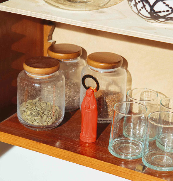 An orange Holy Beer Bottle Opener sits on a wooden shelf with barware and jars of seeds