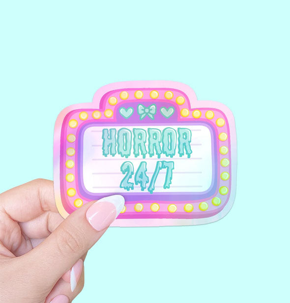 Model's hand holds a sticker designed as a pink and purple movie marquee that says, "Horror 24/7" in drippy blue lettering