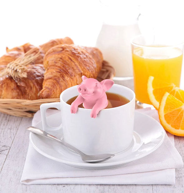Smiling pink pig infuser is placed in a cup tea on a tabletop with pastries, milk pitcher, and orange juice