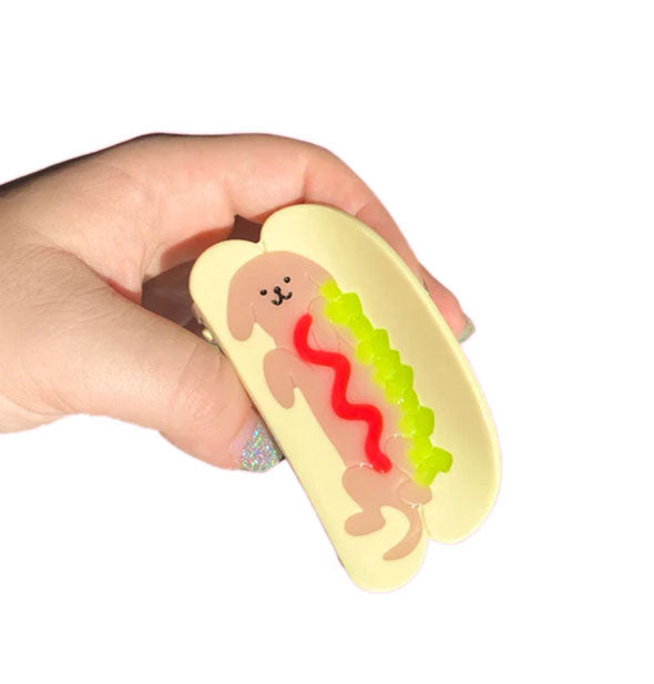 Model's hand holds a light yellow claw clip with design of a dog with ketchup squiggle and relish accent