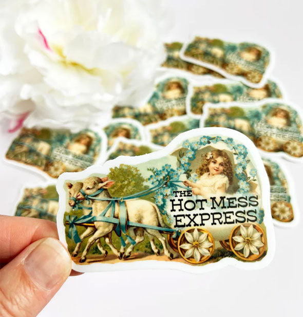 Model's hand holds a Hot Mess Express girl and lambs sticker in front of a pile of others like it in the background staged with a white peony