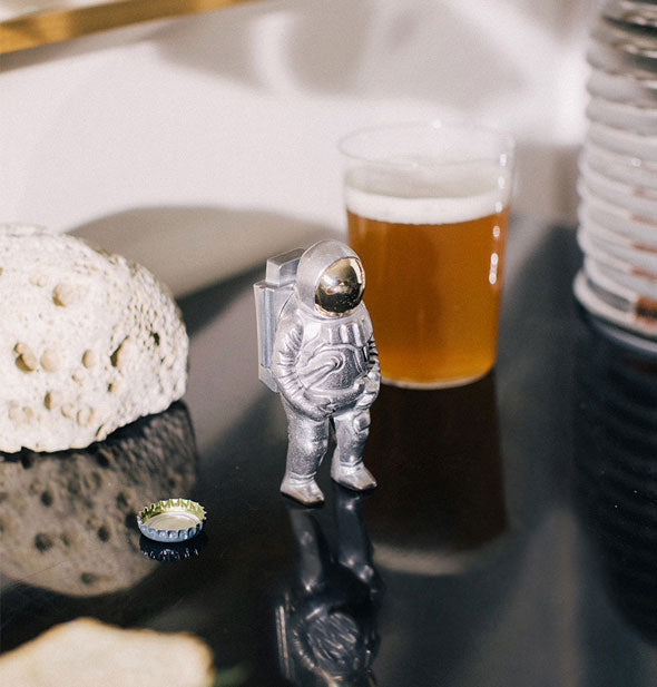 Silver astronaut bottle opener figurine rests on a tabletop next to a bottle cap, glass of beer, and moon rock