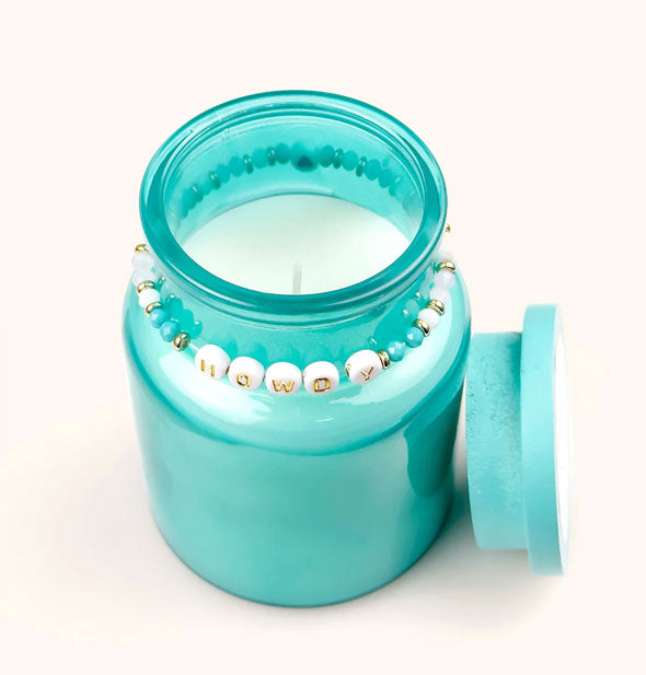 Opened blue-green glass jar candle with beaded "Howdy" bracelet around its neck