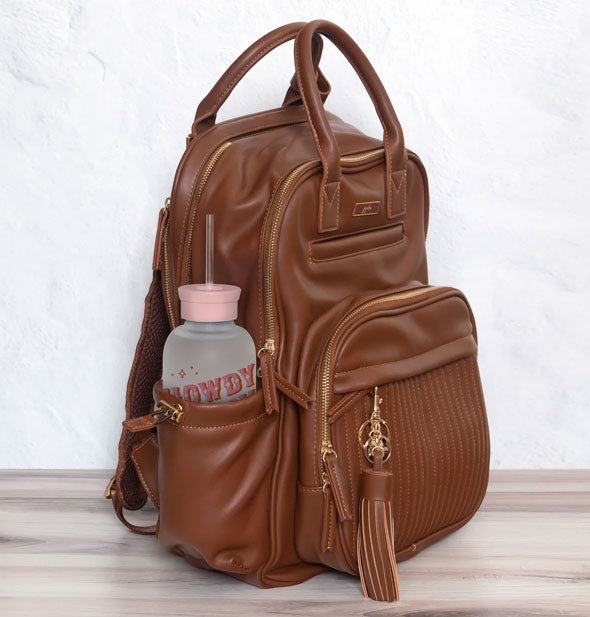 Howdy glass water bottle is tucked into the side pocket of a brown leather backpack