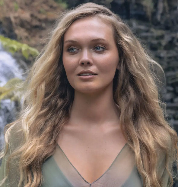 Model with long, blonde hair with a full, wavy, texturized style stands in front of a waterfall backdrop