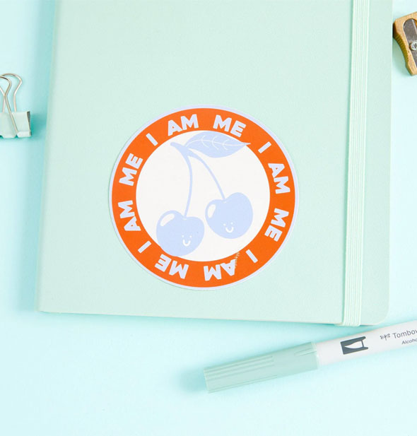 I Am Me Cherries sticker on a light green notebook cover