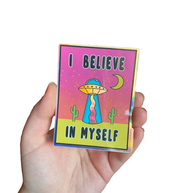 Model's hand holes a rectangular sticker with UFO graphic that says, "I Believe in Myself"