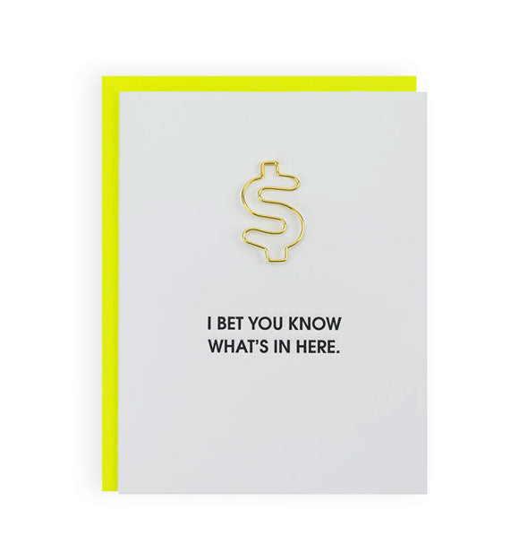 White greeting card with neon yellow envelope partially visible behind it says, "I bet you know what's in here" in minimalist black lettering below a large gold money symbol paper clip